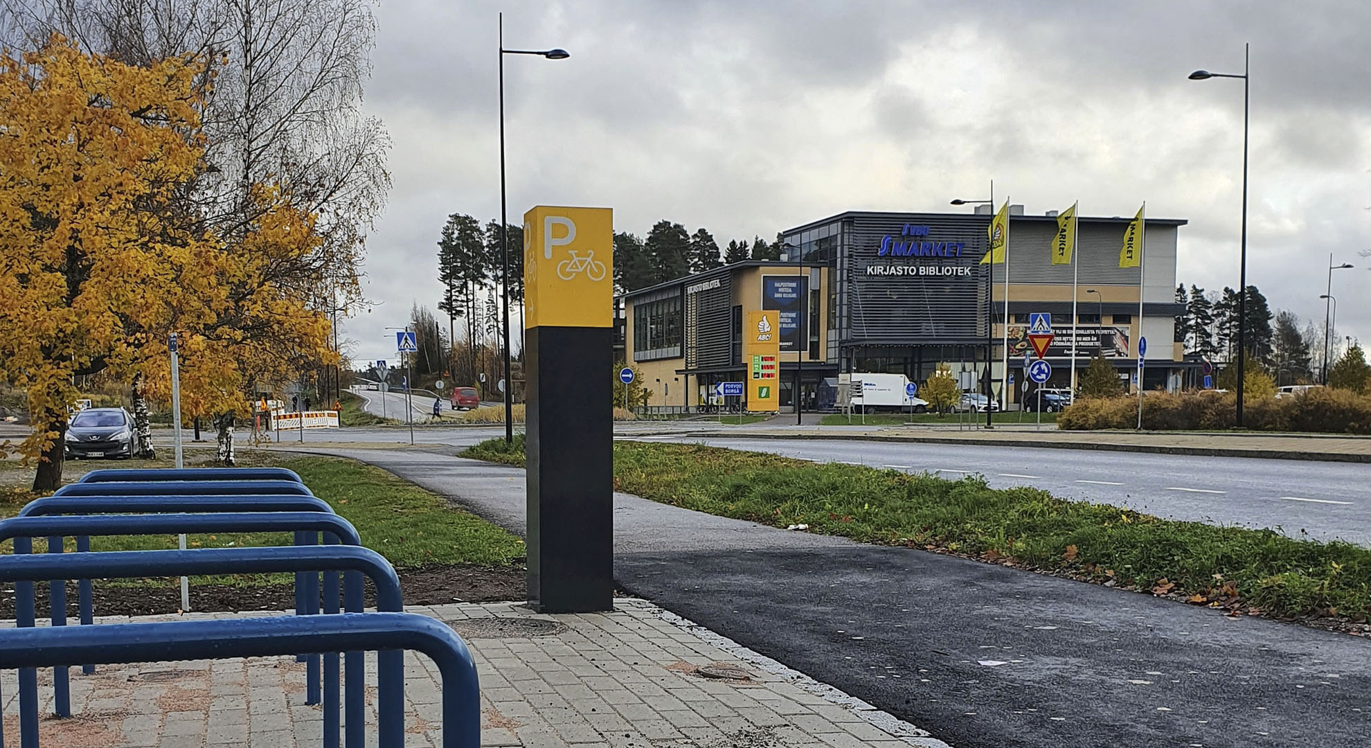 Anslutningsparkeringsplatser för cyklar vid gatan. En skylt med bokstav P och en bild på cykel. Bakom syns Söderkulla centrum.