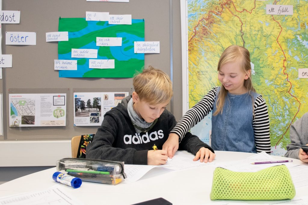 Kaksi lasta pöydän ääressä. Toinen kirjoittaa jotain ja toinen osoittaa paperia sormella. Taustalla seinällä näkyy opetusmateriaaleja ruotsiksi.