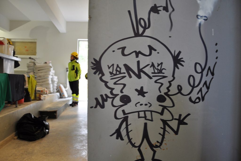 En graffitifigur på väggen och bakom syns byggmaterial och en person klädd i arbetskläder.