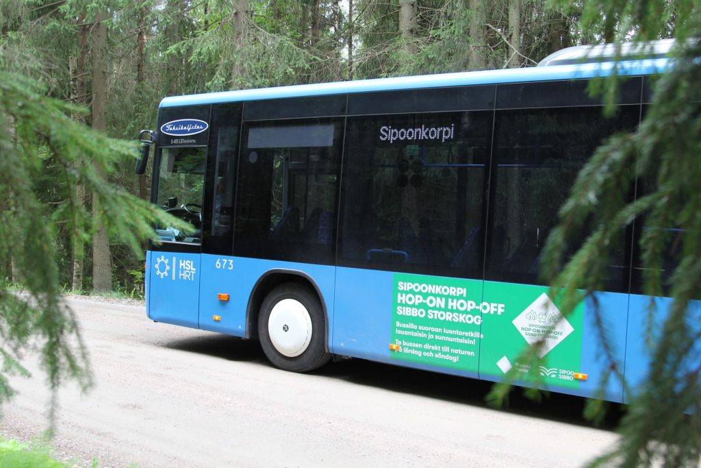 Bussi ajaa puiden reunustamalla tiellä. Bussin kyljessä lukee Sipoonkorpi Hop-on Hop-off.