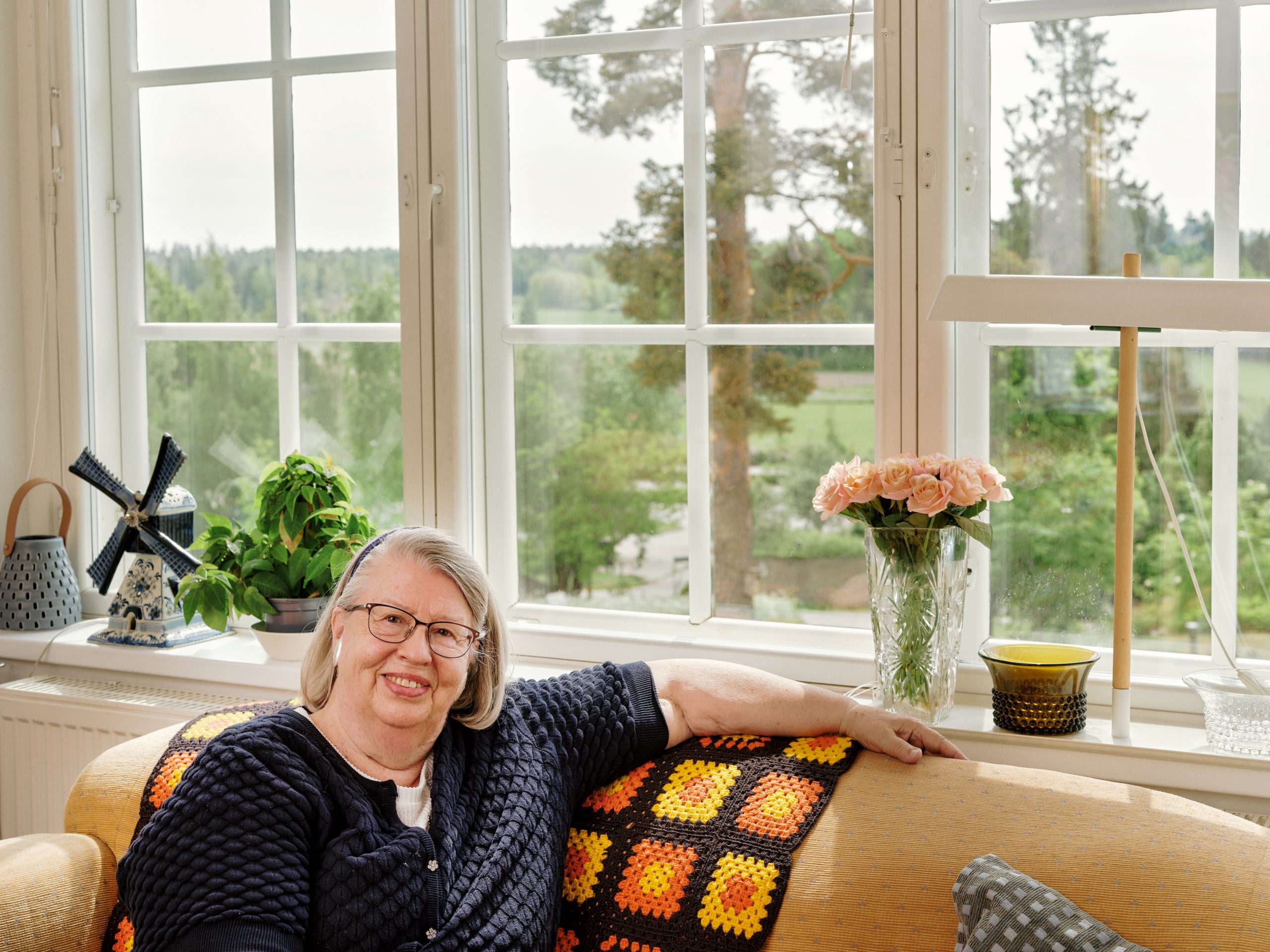 Riitta Nissinen sitter på sin hemmasoffa. Från fönstret bakom henne öppnar sig ett vackert och grönskande landskap.