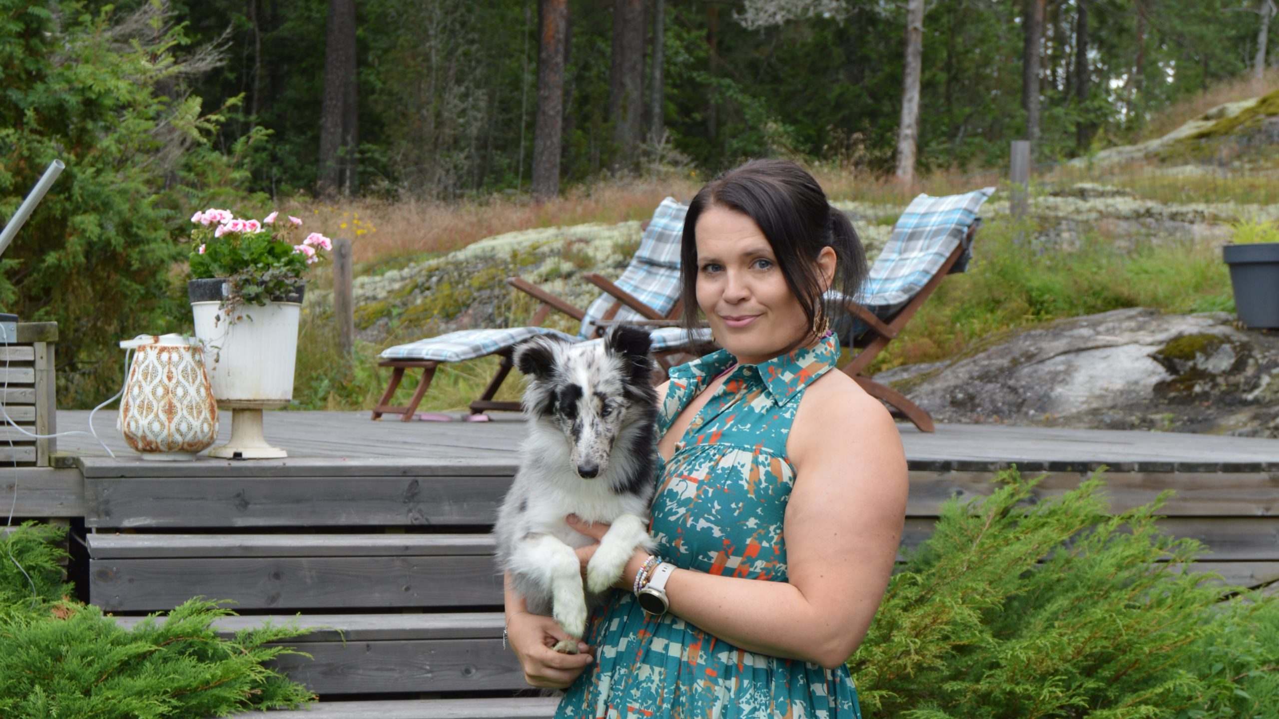 Satu Tuomela står utanför med hunden Lux i famnen. I bakgrunden syns en terrass och skogen.